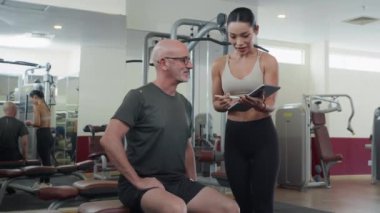 Genç bayan koçun spor kıyafetlerini dijital tabletle çekerken beyaz bir erkeğin spor salonuna ilk gelişinde antrenman planını açıklarken orta uzunlukta bir pozu.