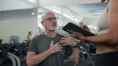 Yetişkin beyaz bir adamın modern spor salonunda dijital tablet kullanarak antrenman planı hakkında kadın koçla konuştuğu fotoğraf.