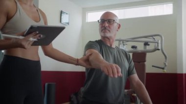 Dijital tablet kullanan kadın eğitmenin düşük açılı görüntüsü. Yetişkin beyaz adamdan modern spor salonundaki özel derste kollarını ve ellerini göstermesini istiyor.