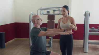 Orta yaşlı Asyalı antrenör, modern fitness merkezinde spor terapisi yoluyla yetişkin beyaz bir adamı desteklerken orta uzunlukta bir çekim.