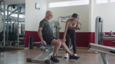Yetişkin bir beyaz adamın bayan Asyalı antrenörle iletişim kurup modern fitness merkezinde rehabilitasyon görürken bacak kaslarını kontrol etmesi.