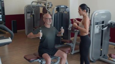 Asyalı bir sporcunun modern spor salonunda spor malzemelerini kullanarak beyaz bir adamın halterlerle egzersiz yapmasına yardım etmesi.