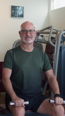 Spor salonunda bayan Asyalı koçla kameraya bakarken elinde dambıl tutan yetişkin beyaz adamın dikey portresi.