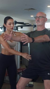 Yetişkin beyaz adamın spor salonundaki bayan Asyalı koçun yardımıyla denge egzersizi yaptığı dikey çekim.