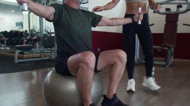 Alçak açı, Asya 'lı kadın antrenörün olgun beyaz bir adamı spor salonunda iki küçük dambılla spor yaparken çekerken görüntüsü.