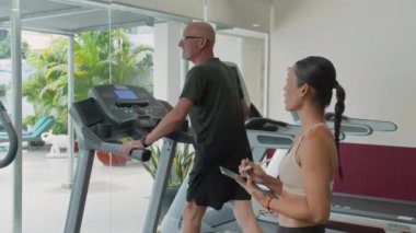 Koşu bandı üzerinde egzersiz yapan olgun beyaz adamın kadın Asyalı koçla spor salonunda dijital tabletle çalışırken geniş açılı yavaş çekimi.