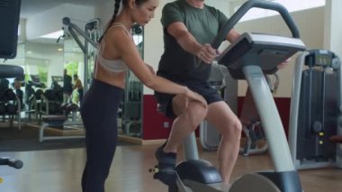 Genç bayan eğitmenin modern fitness kulübünde bisiklete binen yetişkin beyaz adama yardım edişini çek.