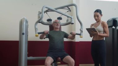 Orta boy kadın çalıştırıcı dijital tablet üzerine notlar alırken erkek yetişkin müşteri spor salonunda makine üzerinde göğüs presi egzersizi yapıyor.