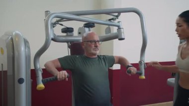 Beyazlı yetişkin bir adamın bireysel fitness dersi sırasında makinede göğüs egzersizi yapmasına yardım eden genç bir kadın eğitmenin el kamerasıyla çekilmiş fotoğrafı.