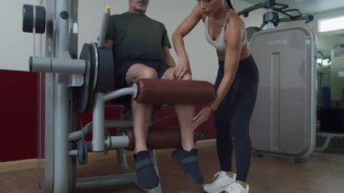 Düşük açı, genç bayan Asyalı koçun spor salonunda spor salonunda yetişkin beyaz adamın bacak çalıştırmasına talimat verirken görüntüsü.