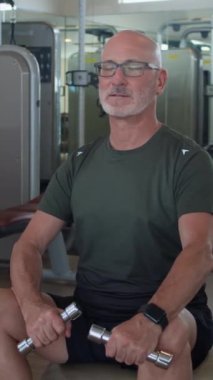 Spor kıyafetleriyle spor salonunda dambıllarla egzersiz yapan ve kameraya bakan yetişkin beyaz adamın dikey portresi.