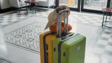 Saman şapka ve seyahat yastığıyla orta boy yeşil ve sarı bavullar şık bir otel lobisinde arka planda zarif mobilyalar.