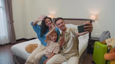 Kafkasyalı mutlu ebeveynlerin ve küçük oğullarının otel odasında yatakta otururken akıllı telefondan konuştuğu görüntüleri yakınlaştır.