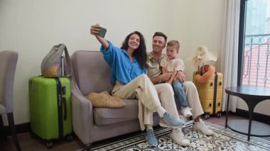 Neşeli Kafkasyalı ebeveynler ve oğullarının kameraya akıllı telefondan poz verip otel odasında selfie çektikleri uzun bir çekim.
