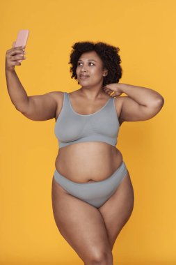 Afro-Amerikalı bir kadın akıllı telefonuyla selfie çekiyor. Gülümserken ve poz verirken sarı bir arkaplanda güvenle durmak