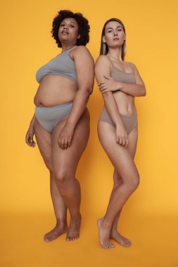Farklı vücut tiplerine sahip iki kadın, özgüvenle parlak arka planda duruyorlar. İkisi de bireyselliği ve kendini kabul etmeyi ifade ediyor.