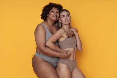 Canlı turuncu arka plana karşı kollarını birbirine dolamış, birbirine yakın duran iki kadın kapsayıcılığı ve kendini sevmeyi vurguluyor.