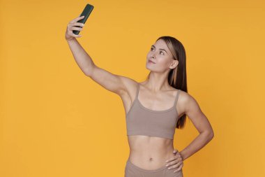 Atletik giyinen kadın sarı arka planda selfie çekiyor. Telefonunu odaklanmış bir şekilde kullanıyor ve kendine güvenerek poz veriyor.