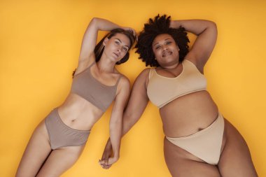 Farklı vücut tiplerine ve etnik kökenlere sahip iki kadın sarı arka planda yatarken tarafsız renk iç çamaşırı giyiyorlar ve özgüven ve güzellik gösteriyorlar.