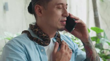 Akıllı telefondan arayan dövmeli bir adamı evinde pitonun boynuna dolanmasıyla etkileşime girerken çek.