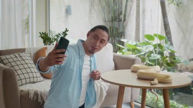 Çok ırklı bir adamın cep telefonunda albino piton yılanıyla selfie çektiği geniş açı evde masada dinleniyor.