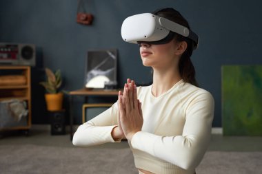 Sıcak ev ortamında sanal gerçeklik kulaklığıyla meditasyon yapan bir kadın. Saksı bitkileri ve modern dekor estetik dokunuş ekleyerek barışçıl bir atmosfer yaratıyor.