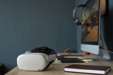 Sanal gerçeklik kulaklığı modern çalışma alanındaki masanın üzerinde, bilgisayar ve defterin yanında. Sahne, günlük çalışma ortamına entegre olan teknolojik gelişmeleri yakalıyor