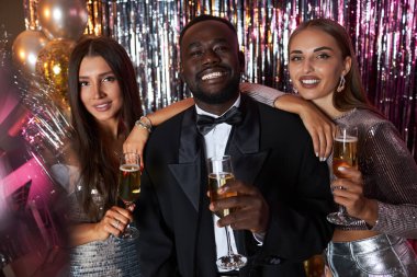 Zarif giyinmiş üç arkadaş, bir etkinliği içkilerle, ışıltılı dekorasyonlarla ve balonlarla kutluyorlar, kameranın önünde içten bir şekilde gülümsüyorlar.