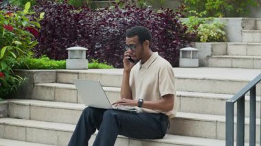 Genç Afro-Amerikan bir adamın akıllı telefondan arayıp şehir merdivenlerinde otururken dizüstü bilgisayar kullanırken orta uzunlukta statik görüntüsü.