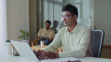 Gözlüklü Hintli erkek çalışanın başlangıç ofisinde kablosuz bilgisayarla rapor verirken orta ölçekli bir fotoğrafı.