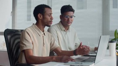 Ofis masasında oturan iki çift ırklı iş arkadaşının kablosuz bilgisayarla ilgili bilgi tartışırken orta boy görüntüsü.