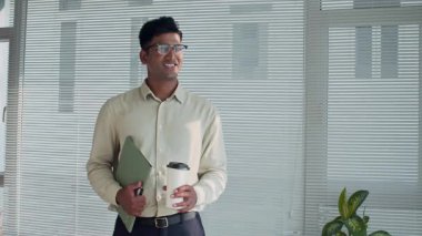 Resmi giyimli ve gözlüklü Hintli bir erkeğin orta boy portresi. Kameraya gülümseyen bir şekilde bakıyor. Elinde kahve bardağı, dosyasında belgeler var.