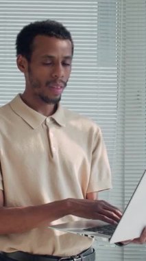 Kablosuz bilgisayarda çalışan ve ofisteki kameraya gülümseyen Afrikalı Amerikalı erkek çalışanın dikey portresi.