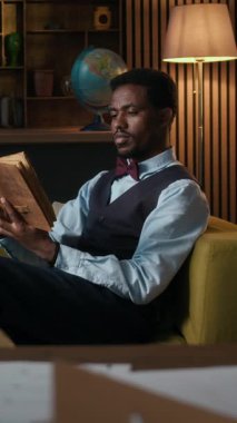 Klasik formalı genç bir adamın kanepede rahat bir şekilde otururken antika kitap okuduğu dikey bir fotoğraf.