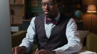 Yelekli, papyonlu ve gözlüklü Afro-Amerikan erkek profesörün merkez ofisteki bilgisayarda çalışmasını çek.
