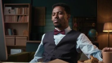 Ofiste bilgisayarın önünde otururken klasik forma giymiş, kurabiye yiyen Afro-Amerikan bir erkek öğretmenin orta boy fotoğrafı.