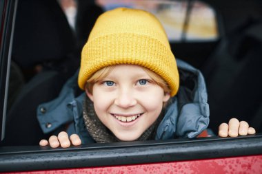 Kış boyunca arabanın camından sarkan sarı bereli gülümseyen çocuğun portresi.