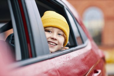 Arabanın camından sarkan sarı bereli mutlu bir çocuk. Parlak bir şekilde gülümsüyor. Arka plan, odak dışı binaları ve çevreyi yansıtıyor.