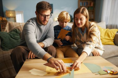 Evde zanaat yapan üç üyeden oluşan aile, masada yaratıcı aktivitelerle meşgul, çocuklu ebeveynler, hepsi de rahat ev unsurlarıyla çevrili projeye odaklanmış.