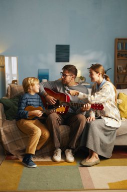 Aile oturma odasında bir araya gelip gitar çalmayı öğreniyor, baba oğlunu eğitiyor, anne çalmaya katılıyor, sıcak aydınlatma ve günlük atmosfer sunuyor.