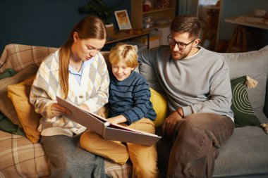 Aile rahat bir kanepede oturup kitap okumaktan keyif alırken, ebeveynler ve çocuklar gülümsüyor ve hikayeyle meşgul oluyorlar, sıcak, kaynaşma anı yaratıyorlar.