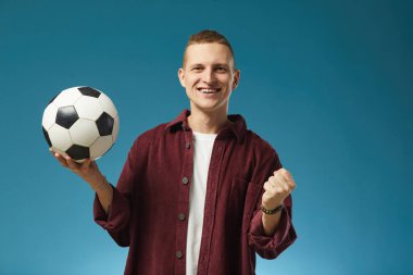 Turkuaz arka planda duran gülümseyen adamın portresi futbol topunu sağ elinde tutarken kestane rengi gömlek giymesi neşe ve coşku göstergesidir.