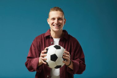 Gülen adam, yüzünde mutlu bir ifadeyle parlak mavi arka planda dururken futbol topunu tutuyor.