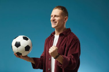 Gülümseyen adam futbol topunu tek eliyle mavi arka planda tutuyor. Kırmızı tişört ve beyaz tişört de dahil olmak üzere günlük kıyafetler giyiyor.