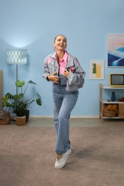 Çağdaş oturma odasının ortasında duran gülümseyen kadın, şık günlük kıyafetler giyiyor, yumuşak ortam ışıklarıyla aydınlatılıyor.