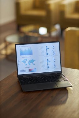 Laptop ahşap bir masada oturuyor ve arka planda bulanık ofis mobilyalarıyla modern bir çalışma alanında iş veri analizlerini temsil eden grafikleri ve grafikleri gösteriyor.