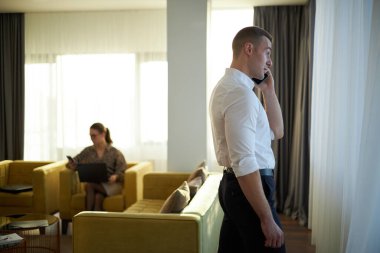 Modern ofiste arka planda dizüstü bilgisayarla çalışan bir iş adamıyla telefonda konuşuyor. Geniş pencereli ve modern mobilyalı parlak oda