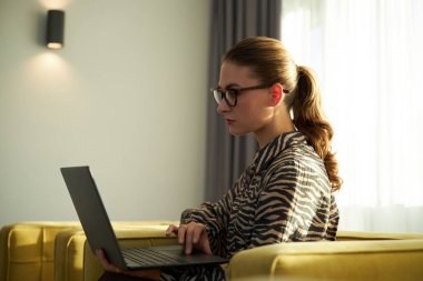 Sarı koltukta oturan, dizüstü bilgisayarla çalışan, penceresinden yansıyan doğal ışık ve gözlük takan, modern bir oturma odasında çalışan bir kadın.