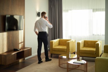 Modern bir otel odasında akıllı telefondan konuşan ve geniş bir pencereye bakan bir iş adamı. Odada çağdaş mobilyalar ve bol miktarda doğal ışık bulunuyor.