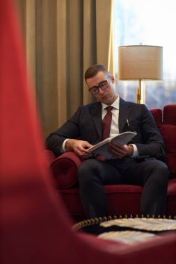 Genç işadamı kırmızı kanepede resmi giyinmiş, gözlüklü belge okuyor, perde arkası ve masa lambasıyla şık ofis alanına odaklanmış görünüyor.
